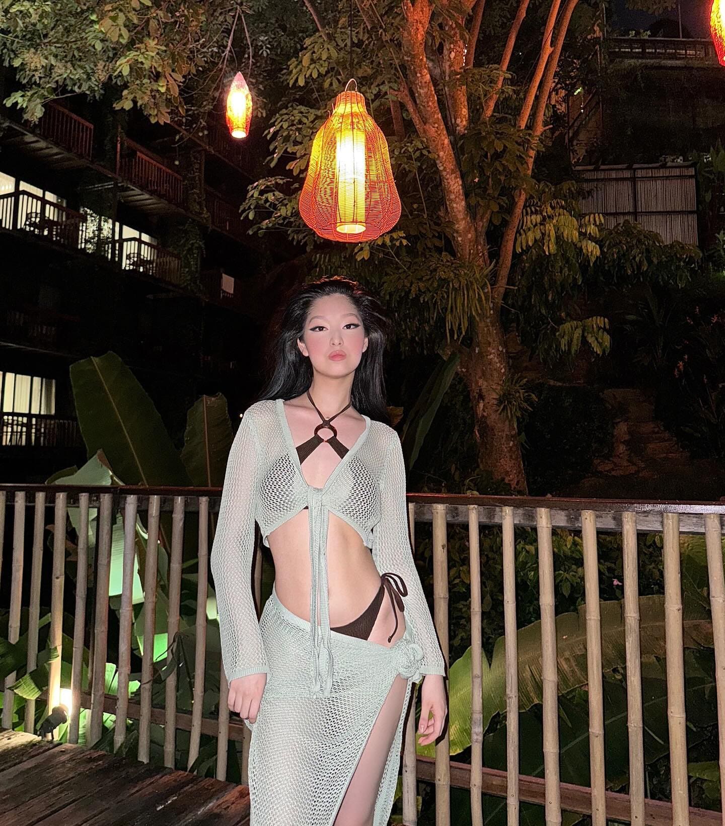 深色长发女性身穿深棕色比基尼，外搭浅绿色透视镂空长袖上衣和高开叉长裙，夜晚站在有热带植物和灯笼的木质阳台上。