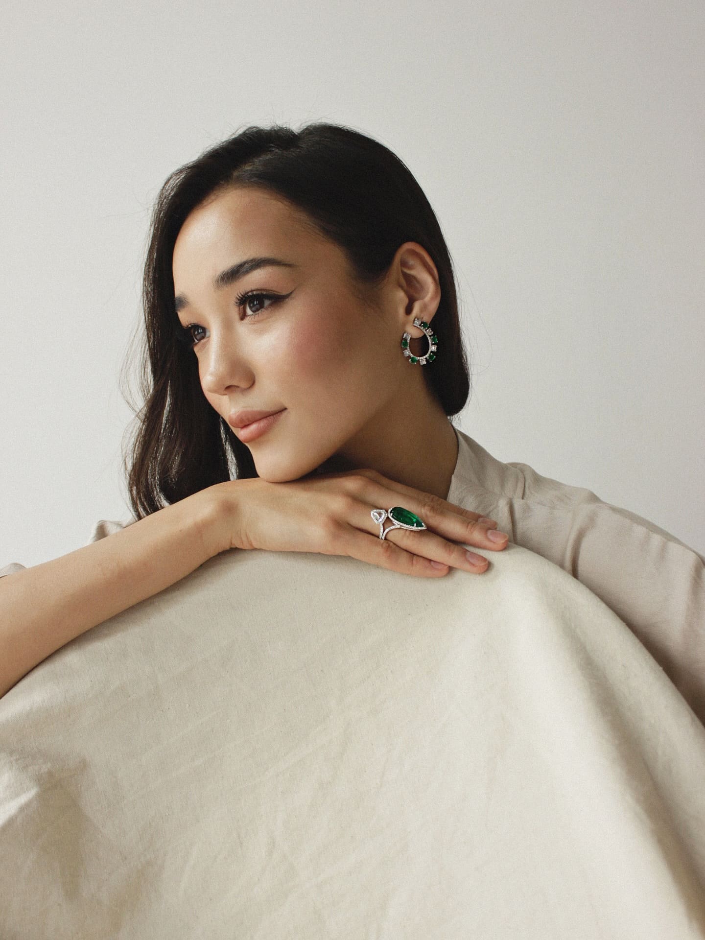 Bayan rests her chin on her hand, wearing a high-necked beige garment and large green gemstone jewelry against a plain light background.