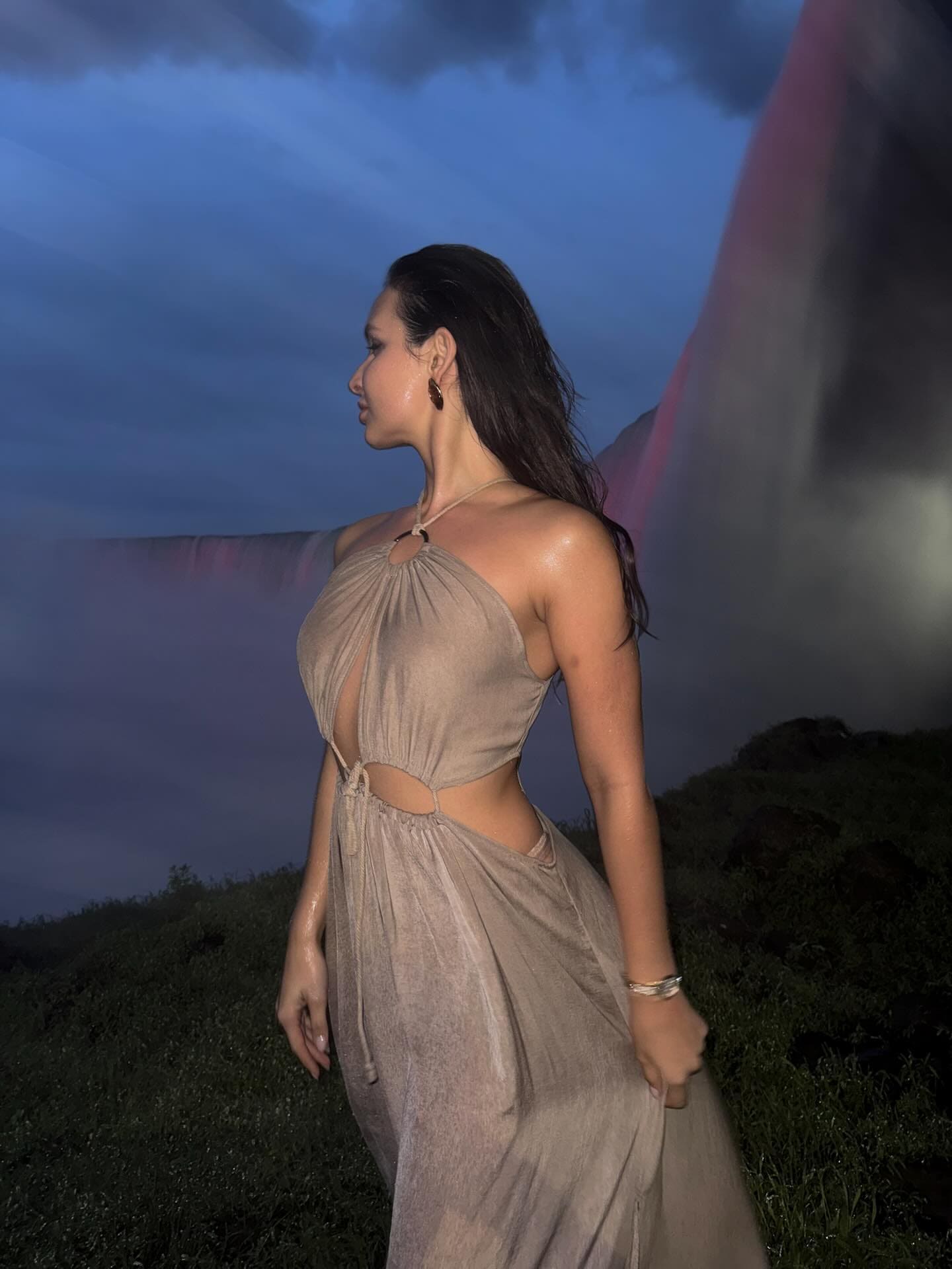 A woman stands in profile against a twilight sky in a beige halter maxi dress with a plunging neckline and sheer skirt.