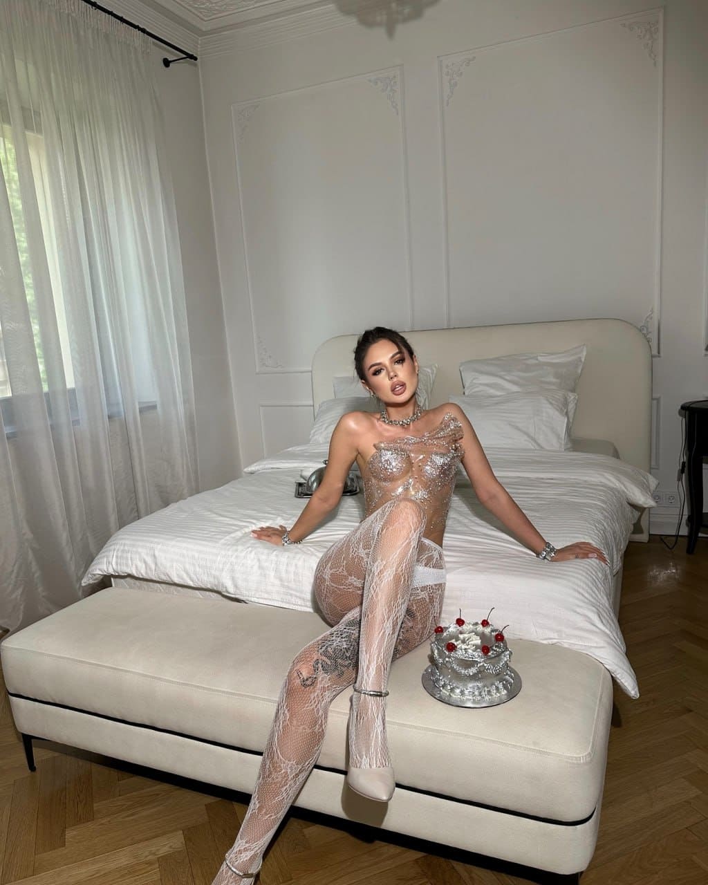 A woman sits on the edge of a bed, wearing a sheer embellished bodysuit and patterned tights, posing with one leg crossed next to a small cake.