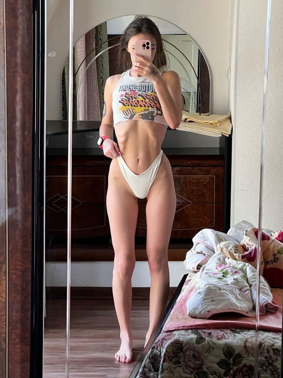 A mirror selfie shows a woman in a graphic white crop top and briefs, standing barefoot near a wooden dresser and arched mirror.
