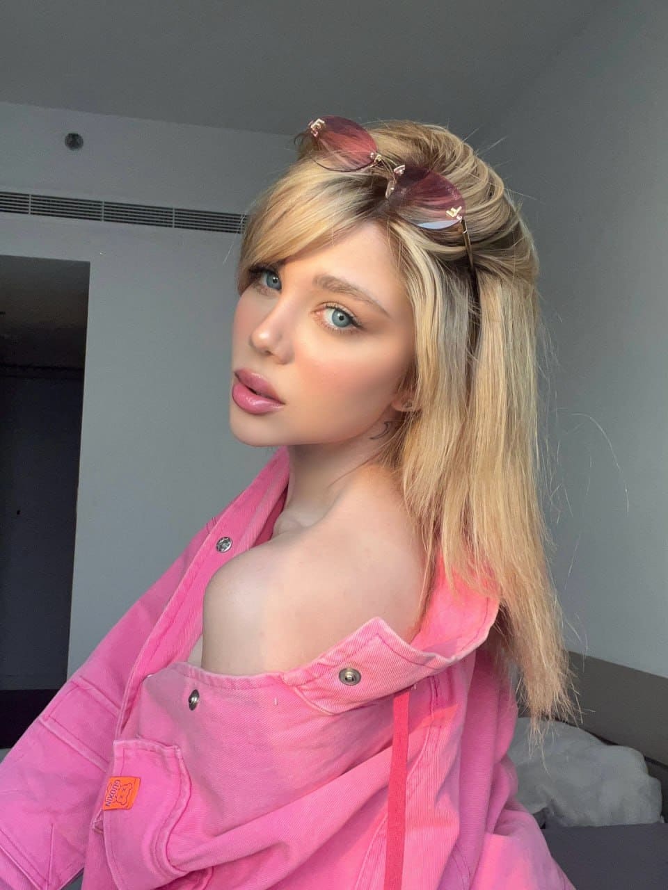 Marianne looks over her shoulder in a pink denim jacket draped off one shoulder, standing against a plain grey wall.