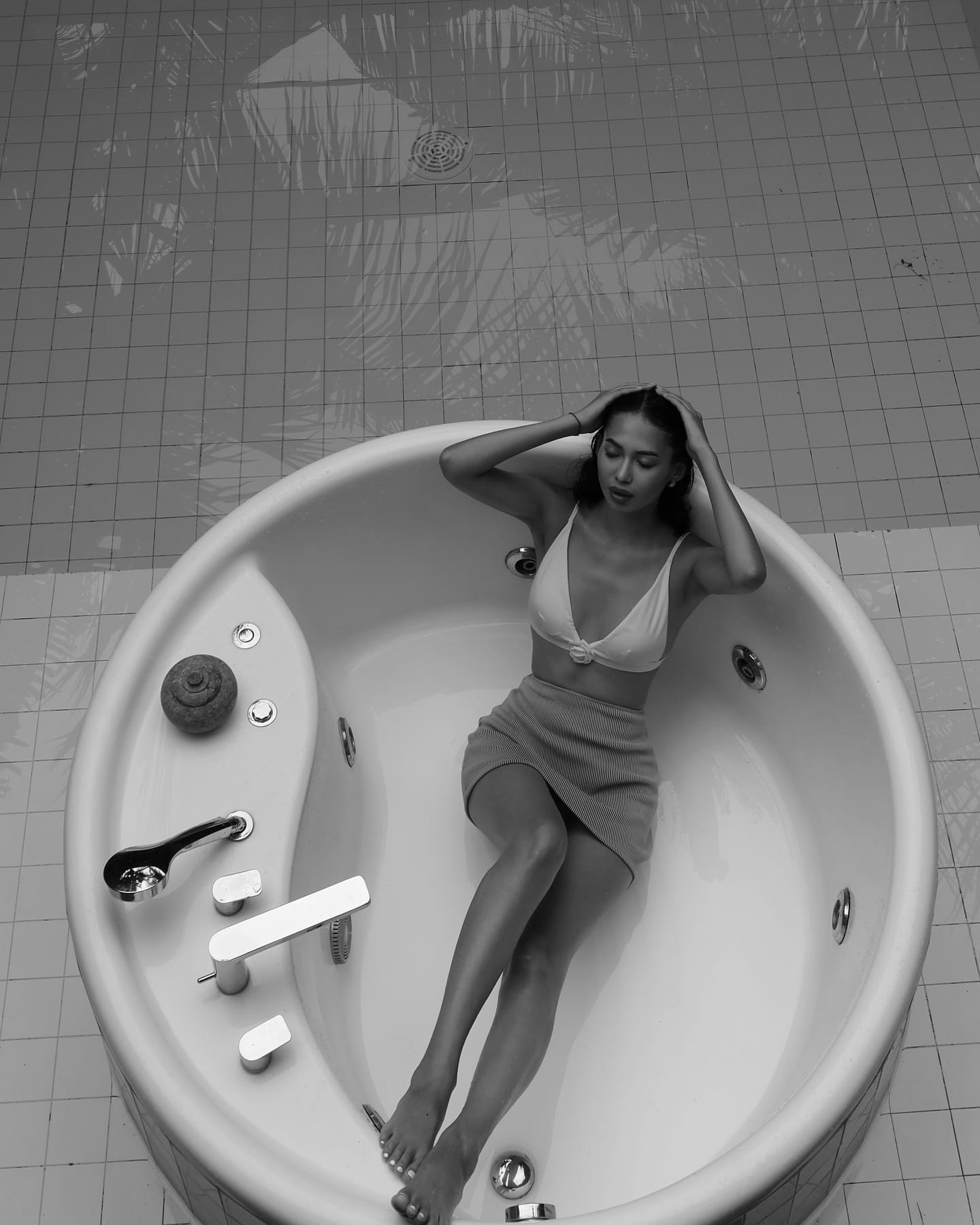 A woman sits in a white circular bathtub, wearing a light bra top and short skirt, hands on head, legs extended.