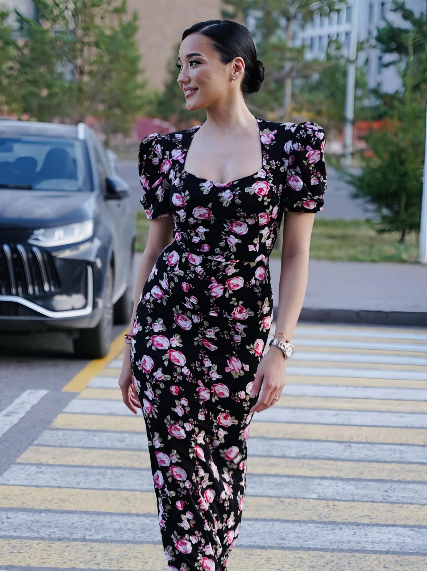 A woman in a black midi dress with pink floral print and puffed sleeves stands on a crosswalk, a dark SUV visible in the background.