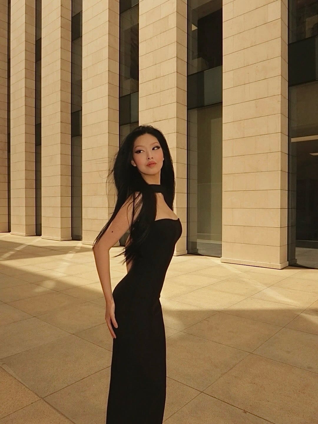 深色长发女性侧身站立，身穿黑色抹胸长礼服并佩戴同色颈圈，背景为高大的米色石柱。