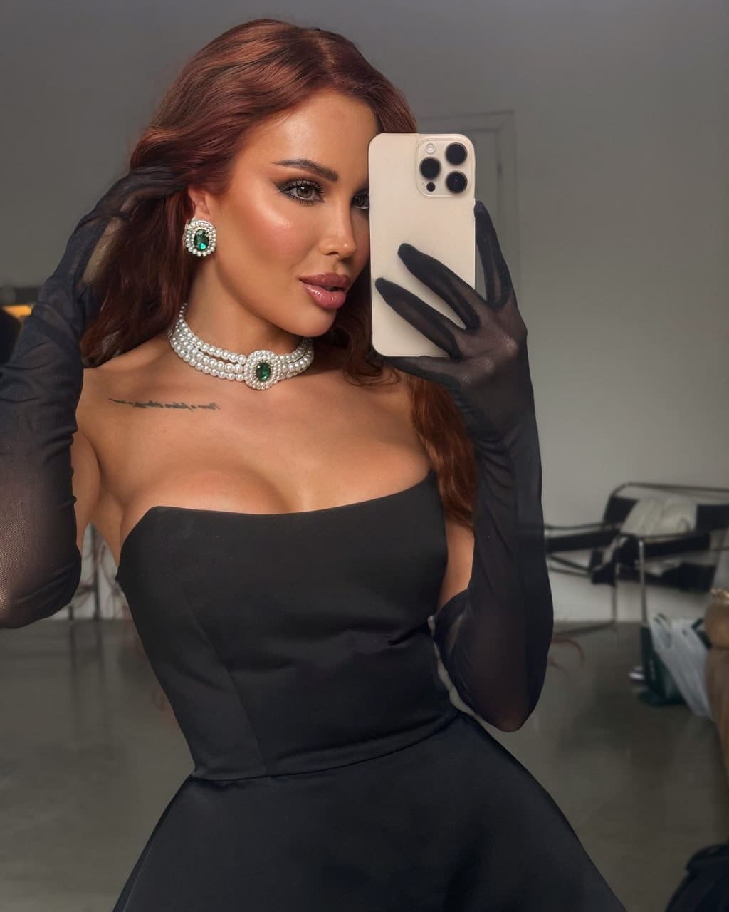 A woman takes a mirror selfie in a black strapless dress and sheer opera gloves, wearing a pearl choker and earrings.