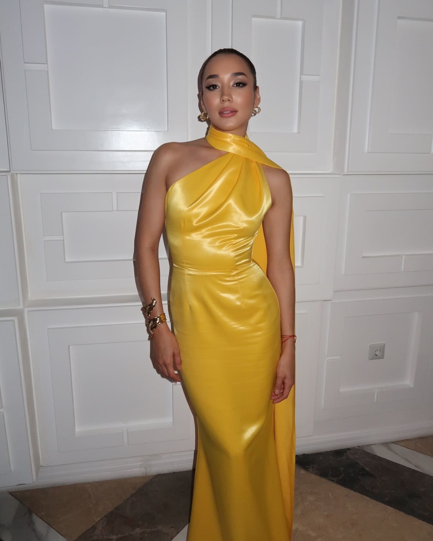 A woman in a floor-length yellow satin gown with a one-shoulder design and gold jewelry stands against a white paneled wall.