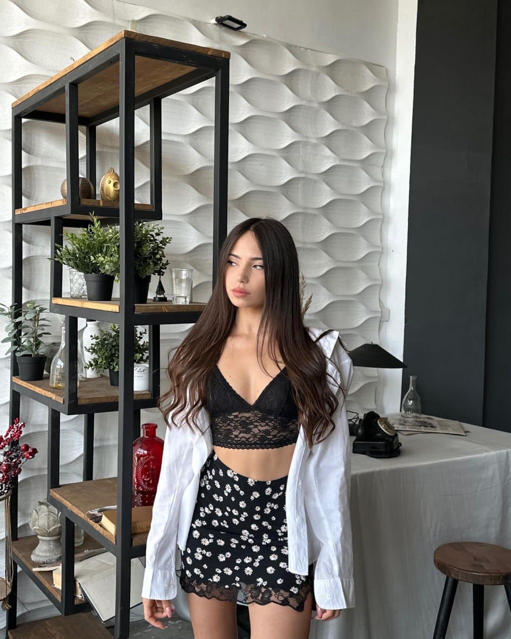 Josephine stands indoors in a black lace bralette, floral mini skirt, and open white shirt before a metal shelf and textured wall.