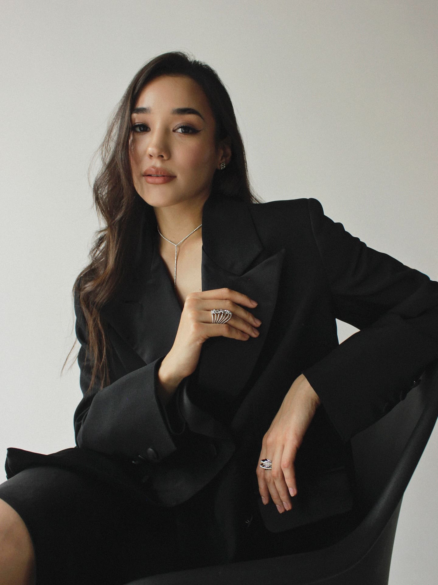 A woman sits in a black chair wearing an oversized black blazer and silver lariat necklace, one hand resting on her chest.