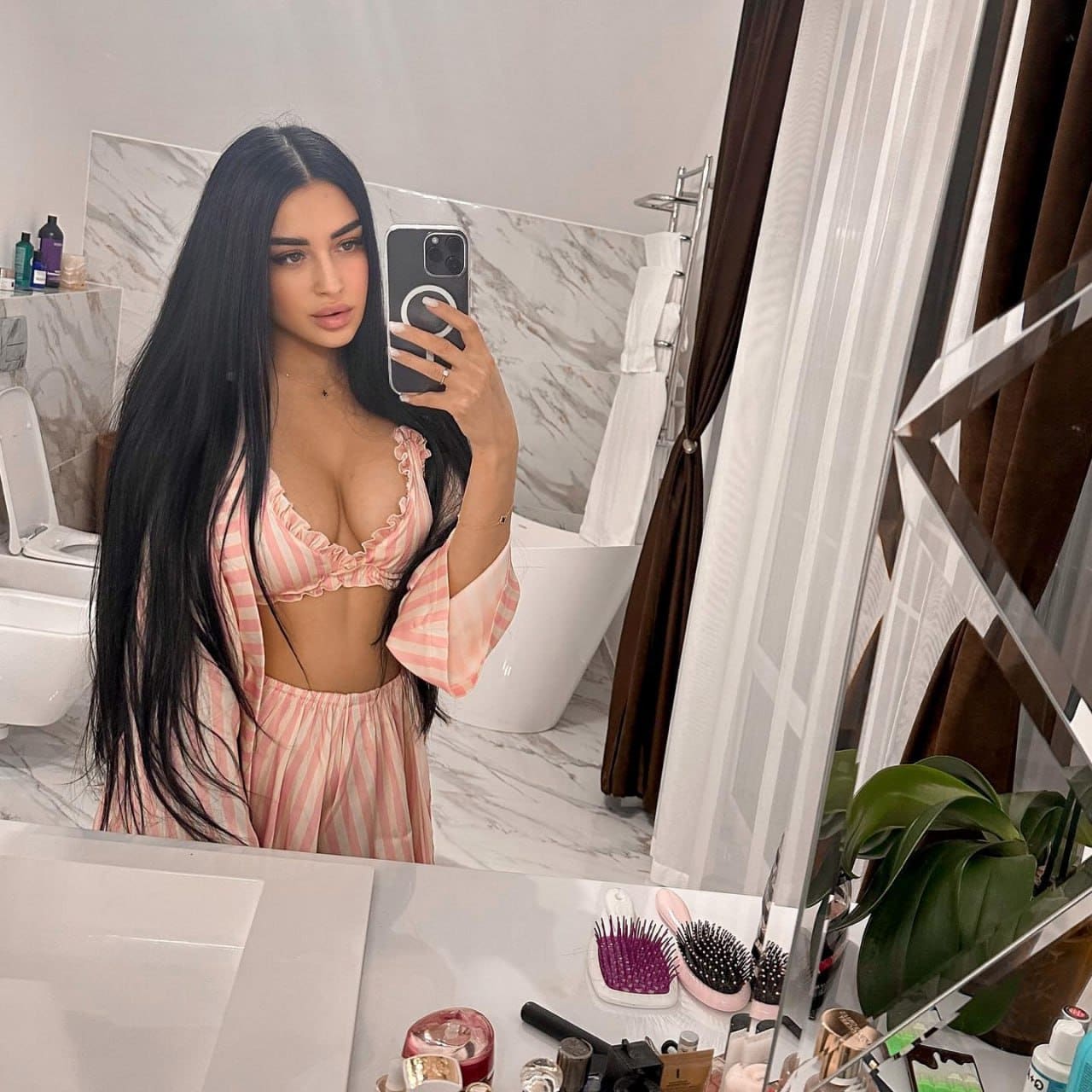 A mirror selfie in a bathroom features a woman wearing a pink and white striped ruffled bra top and matching bottoms with an open midriff.