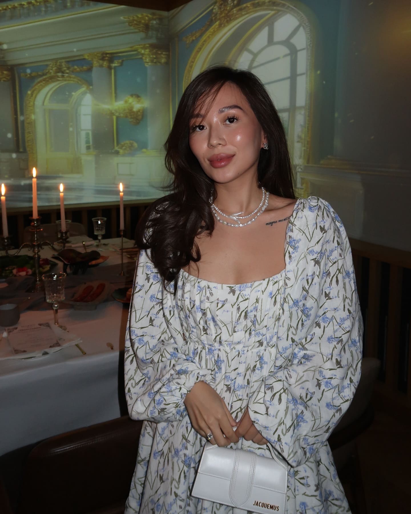 She stands indoors in a white dress with blue floral print, square neckline, and puffed sleeves, wearing a pearl necklace and holding a white handbag.