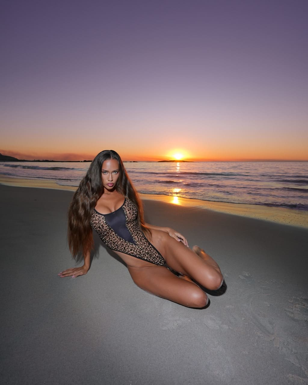 Kneeling on a sandy beach at sunset in a high-cut leopard-print bodysuit.