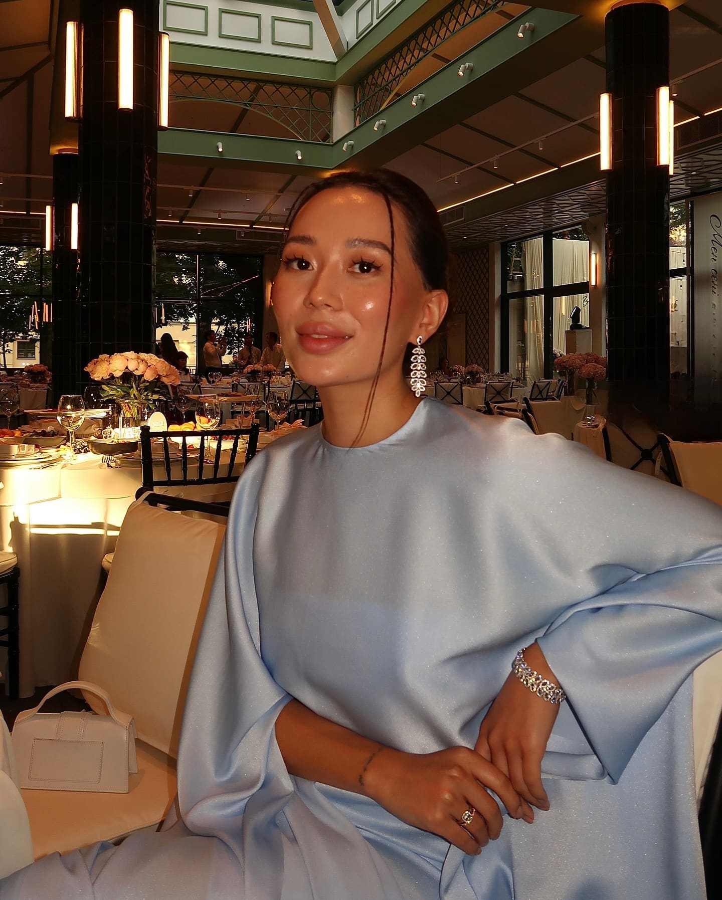 Seated in a restaurant, she wears a high-necked light blue satin gown with voluminous sleeves, accessorized with dangling earrings and a bracelet.