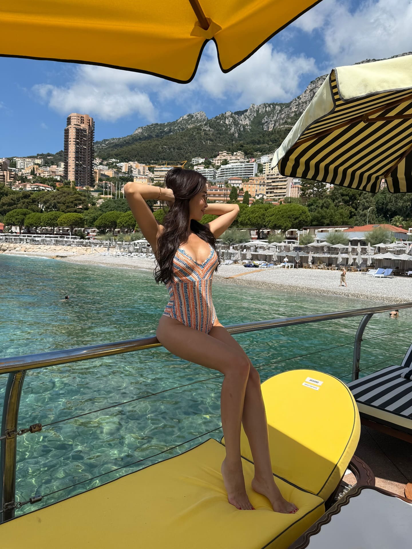 A woman in a patterned one-piece swimsuit sits on a yellow lounge chair, hands behind her head, against a coastal backdrop of mountains.