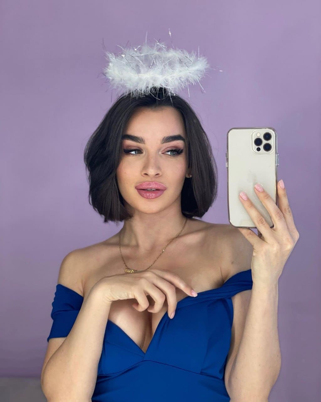 Estelle takes a mirror selfie in a blue off-the-shoulder dress and feather halo headband against a purple background.
