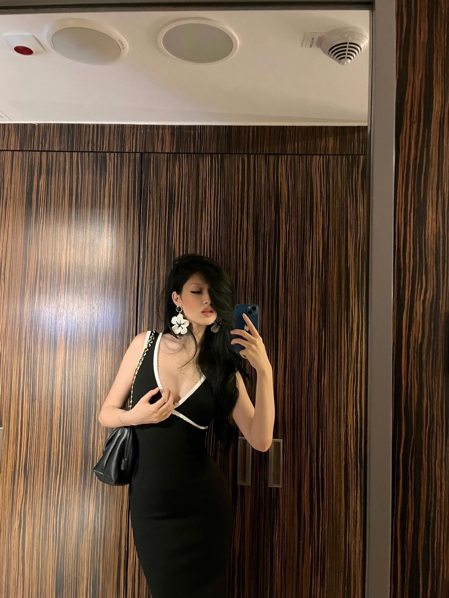 深色长发女性在木饰面墙前对镜自拍，身穿黑色深V领无袖连衣裙，领口有白色滚边，一手持手机，肩背黑色手提包。