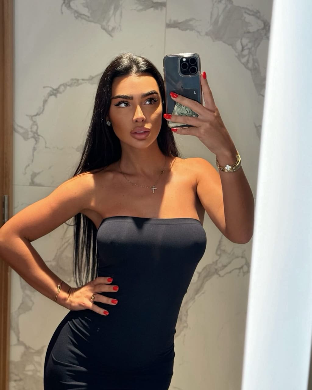 Ameliya takes a mirror selfie in a strapless black dress, hand on hip, against a marble-tiled wall.