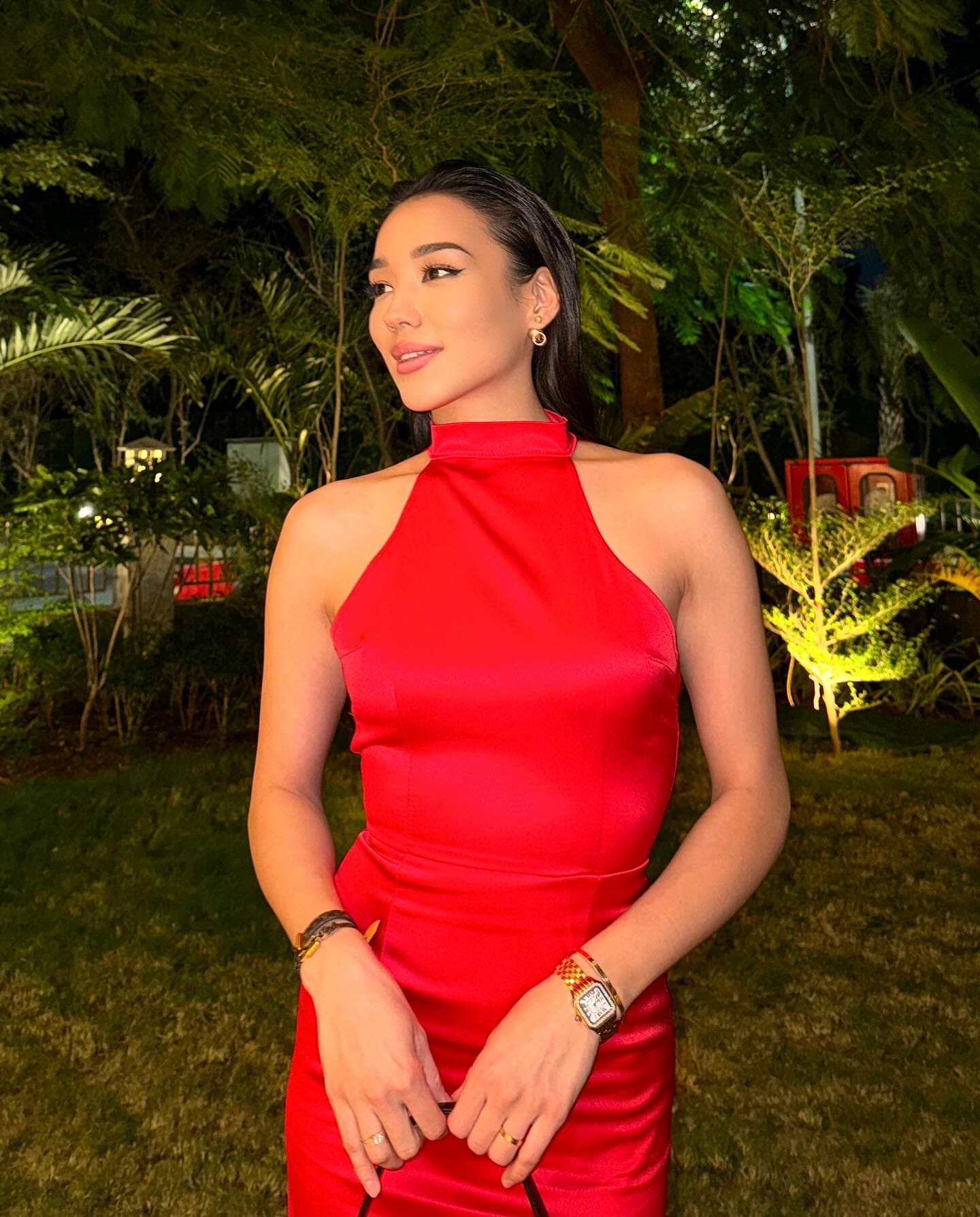 A woman with slicked-back hair stands outdoors at night in a fitted red satin halter dress, hands clasped in front of her waist.