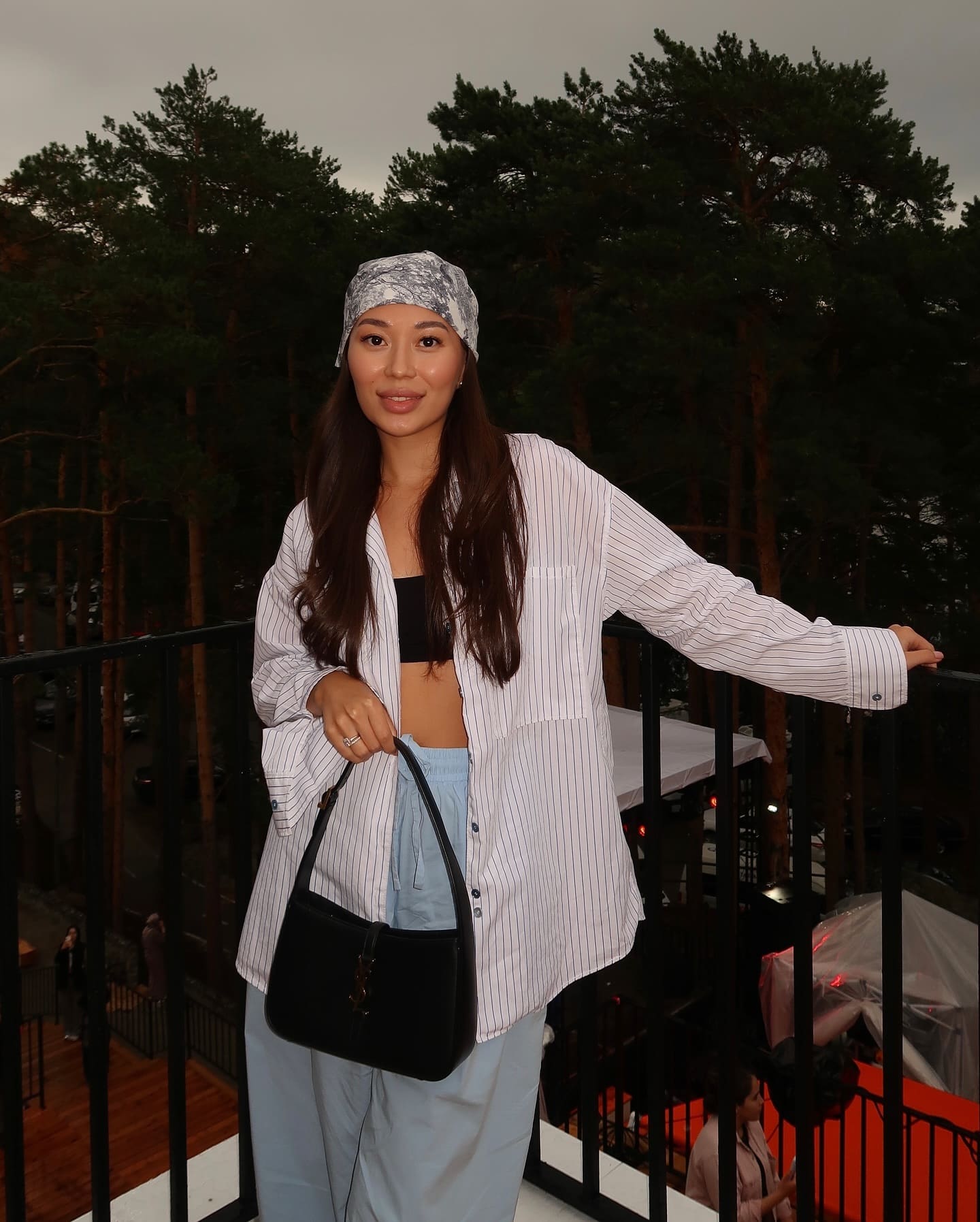On a balcony, she wears a patterned headscarf, an open white pinstripe shirt over a black bandeau top, and light blue trousers, holding a black handbag.