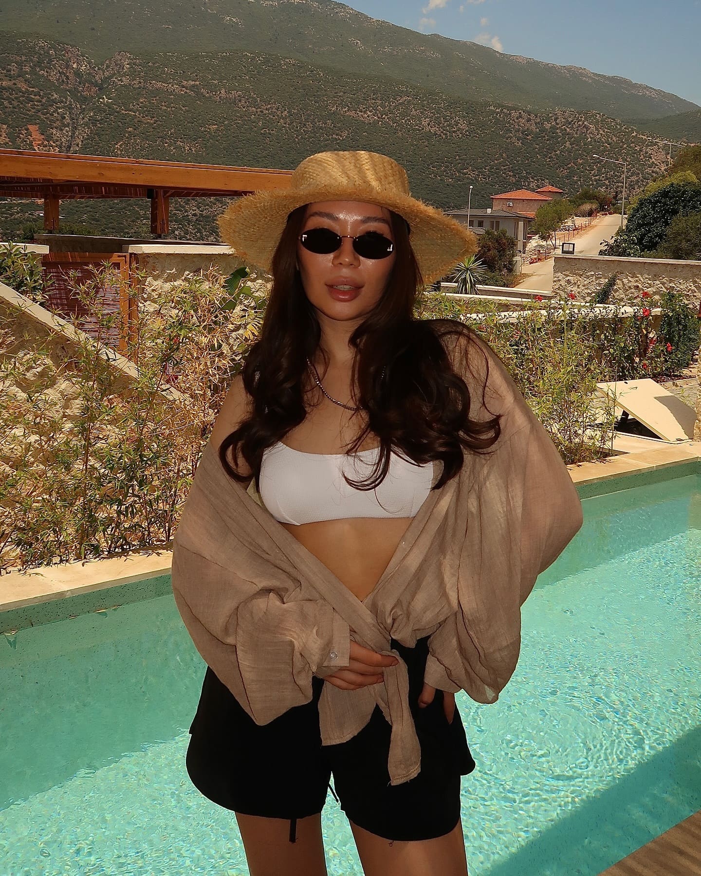 By a swimming pool, she wears a wide-brimmed straw hat, sunglasses, white bandeau top, black shorts, and a loose beige shirt draped off her shoulders.
