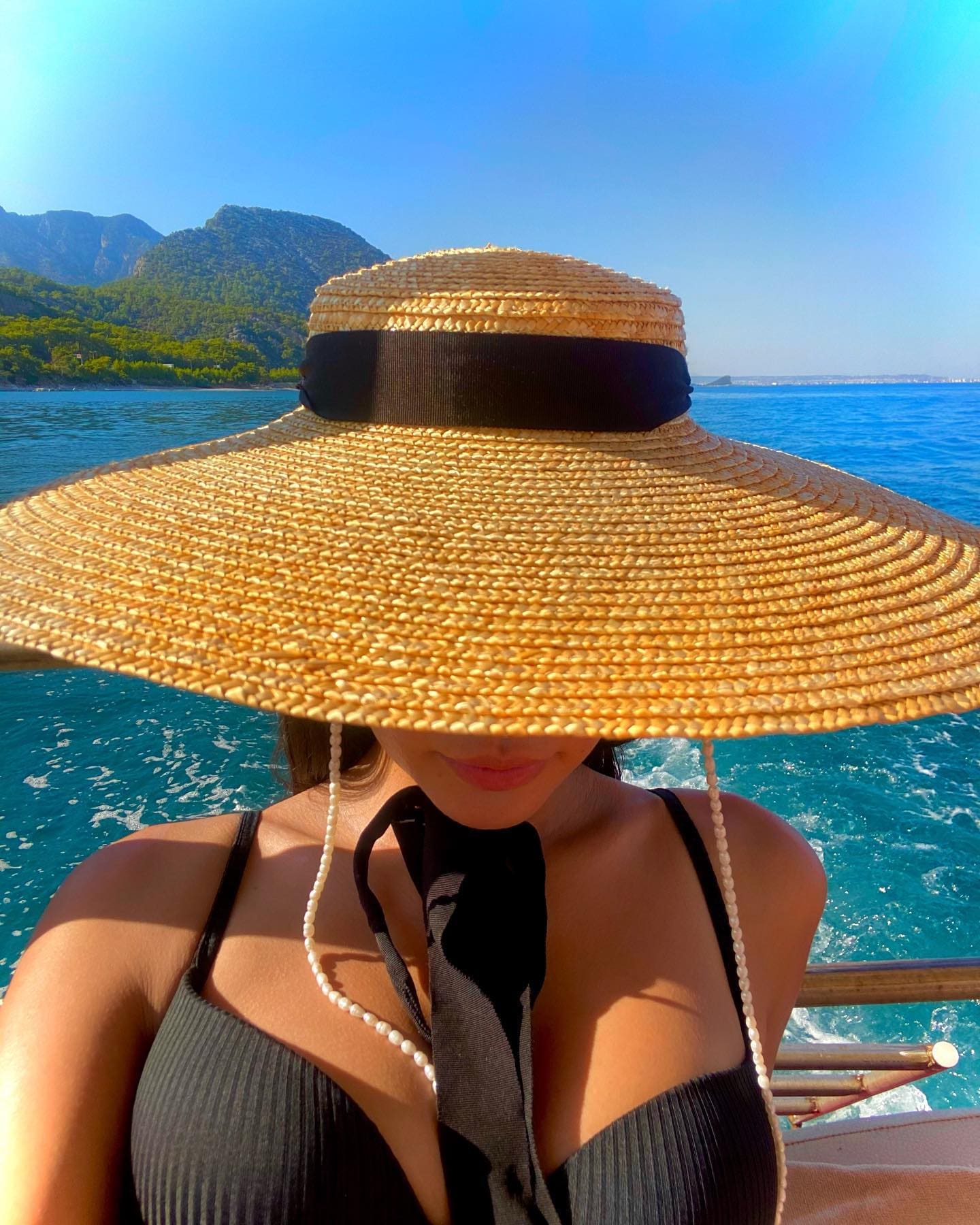 A woman wearing a wide-brimmed straw hat and black ribbed top poses on a boat with blue sea and mountains in the background.