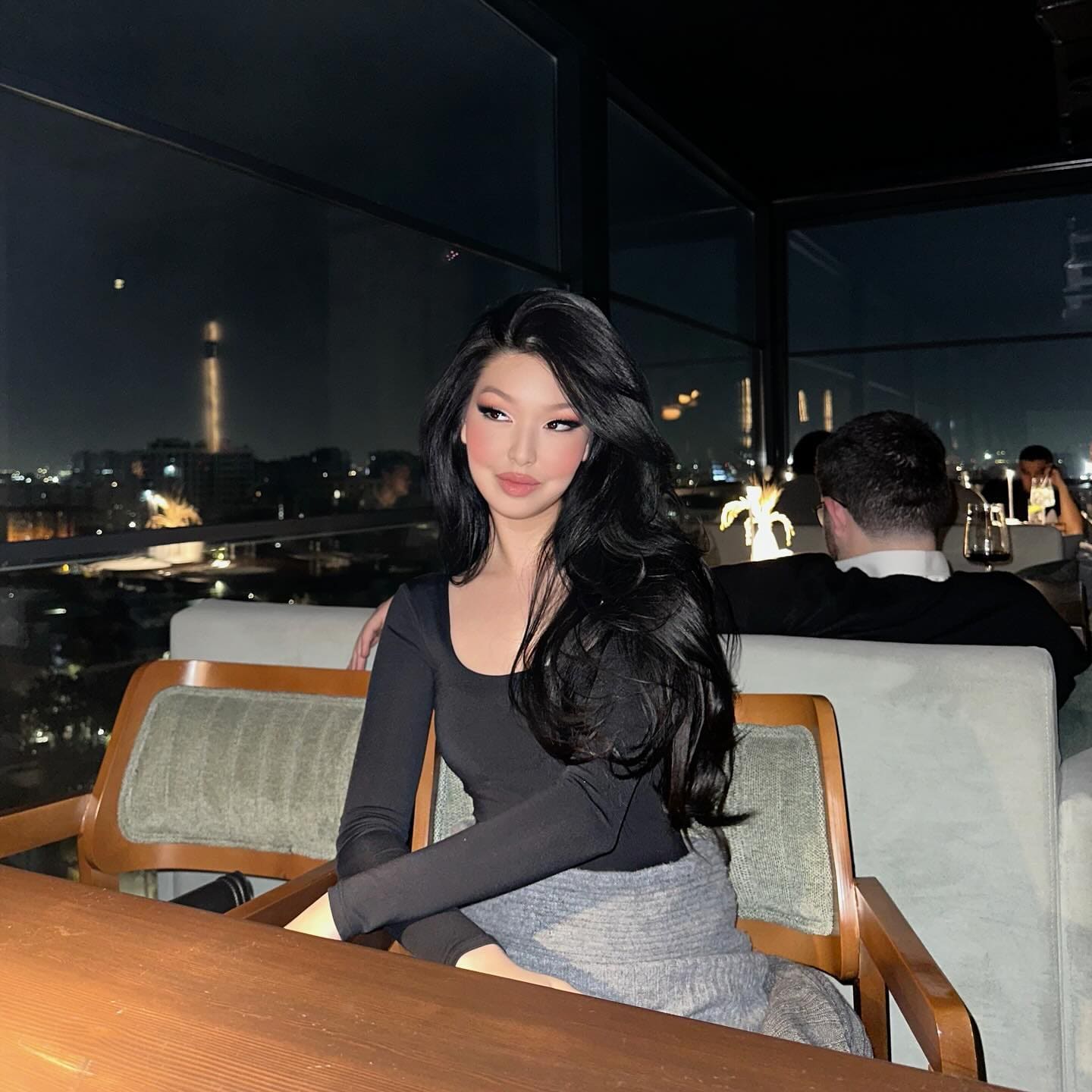 深色长发女性身穿黑色长袖圆领上衣和灰色裙子，坐在灯光昏暗的休息区桌边，身后窗户可见城市天际线。