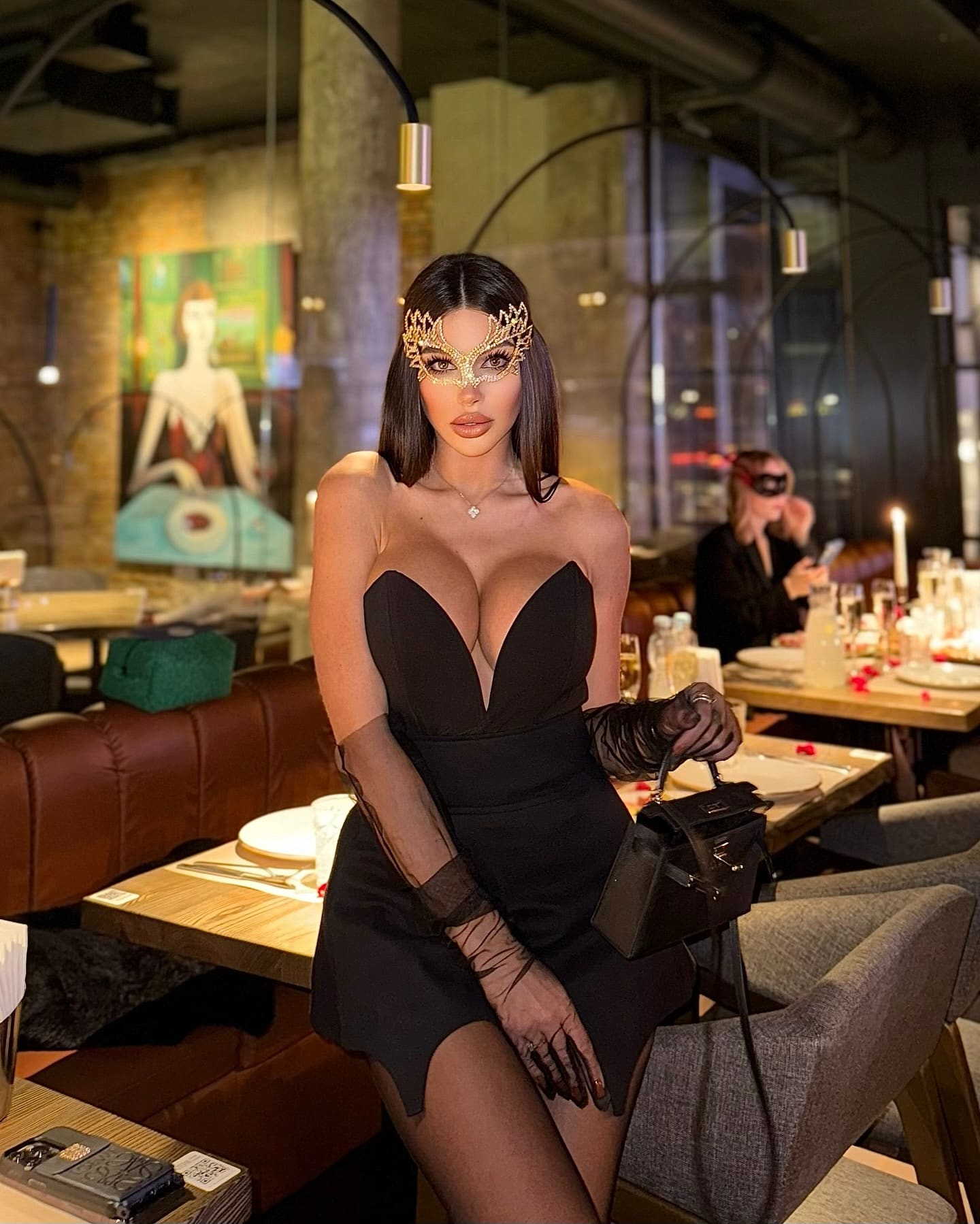 Woman in a restaurant wearing a black strapless dress, opera gloves, and a gold eye mask, holding a black handbag.