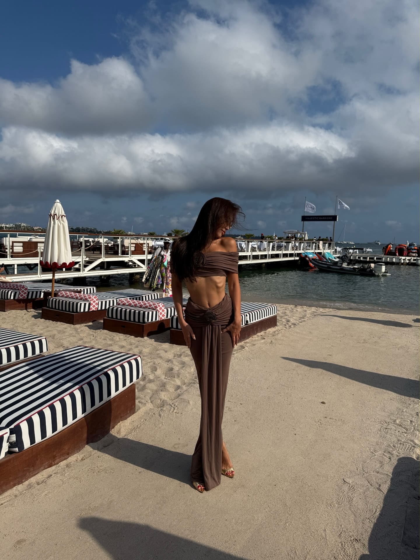 A woman stands on a sandy beach in a brown off-shoulder top and draped skirt, with striped lounge chairs and a pier visible in the background.