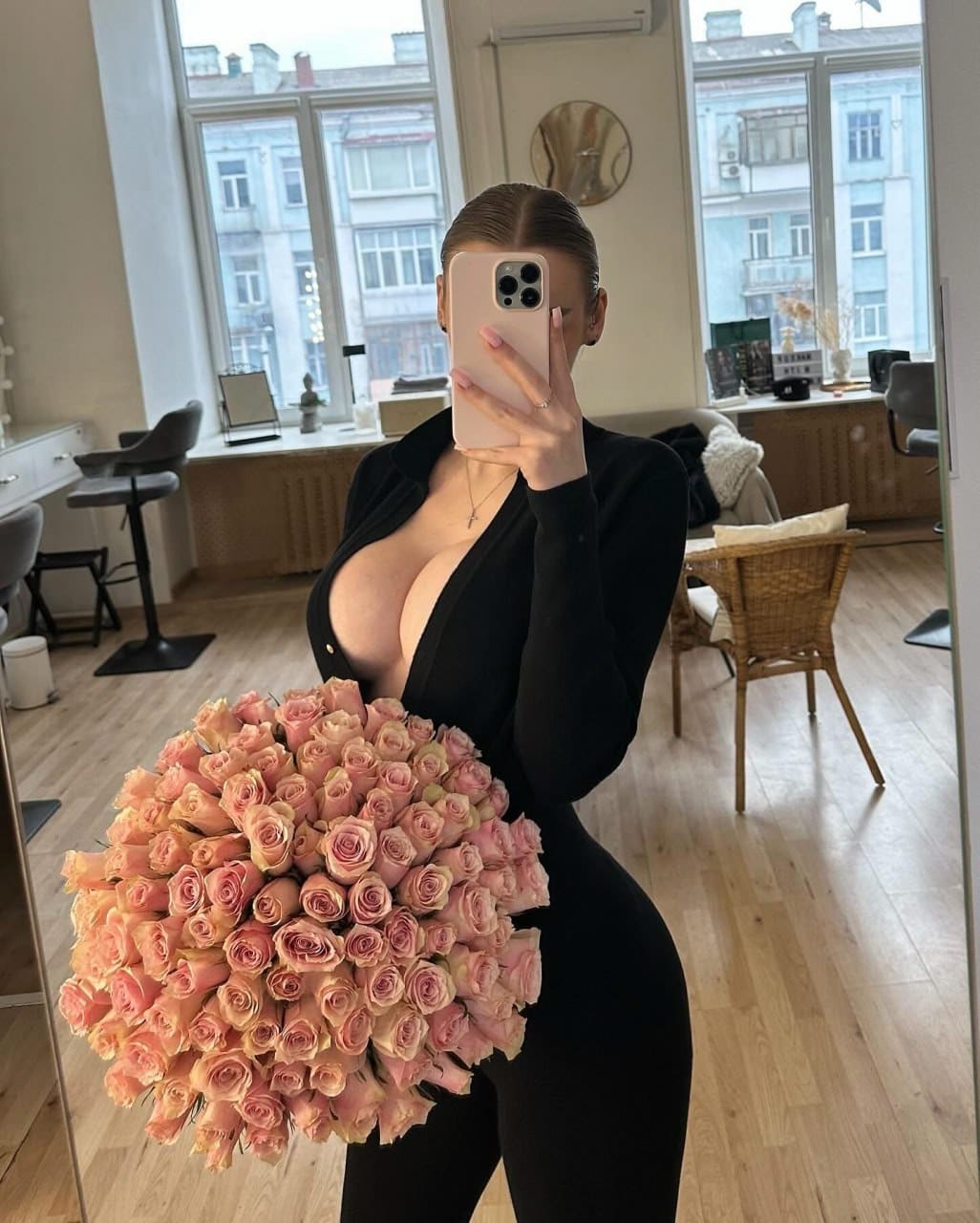 A mirror selfie shows a woman with slicked-back hair holding a phone and pink roses, wearing a tight black bodysuit with a deep plunging neckline.