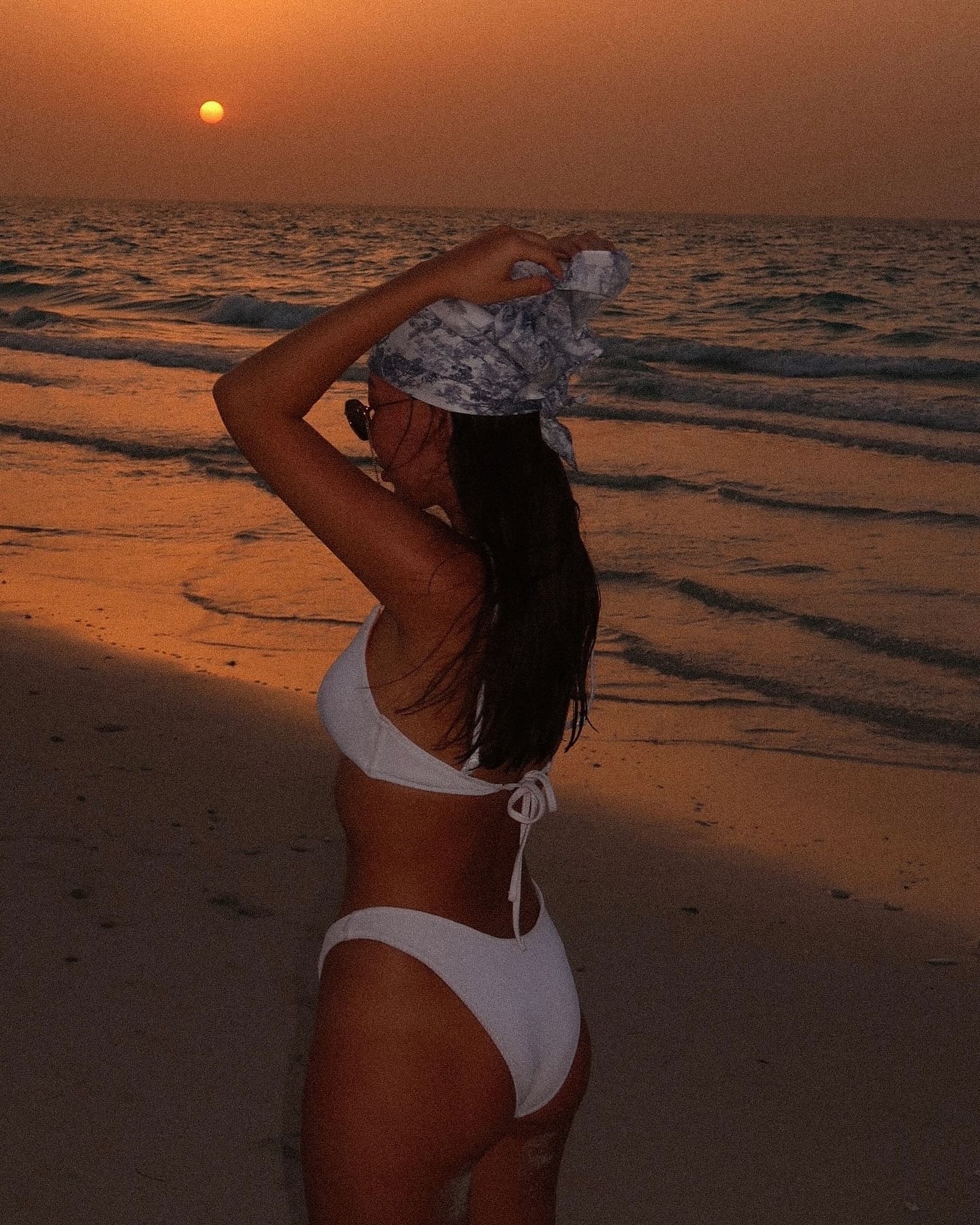 On a beach at sunset, she stands in profile wearing a white string bikini and patterned headscarf, one arm raised to hold the scarf while facing the ocean.